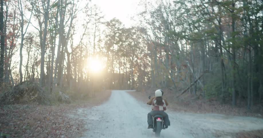 Two people ride a motorcycle through the forest in autumn as the sun nears the horizon