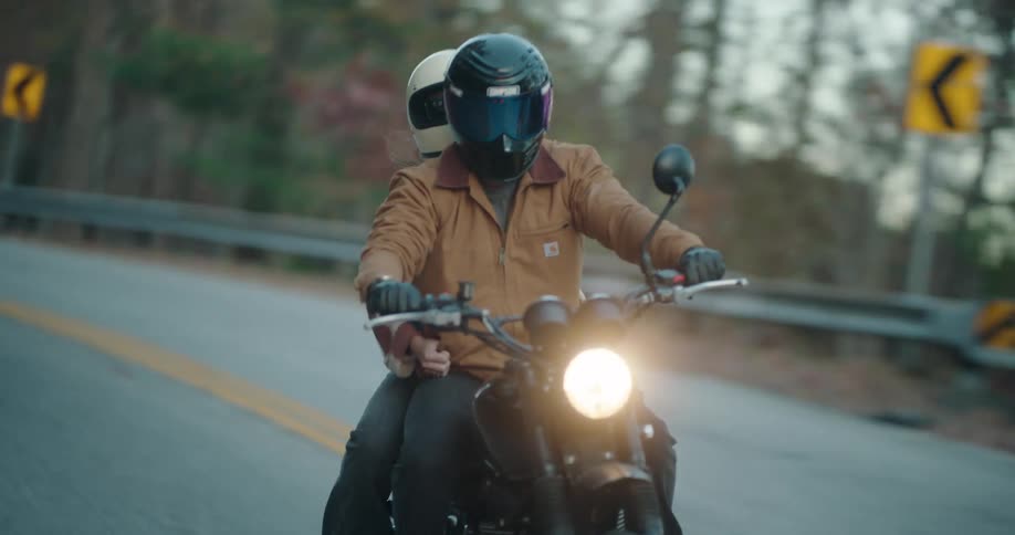 Headlight glows on a motorcycle traveling with a passenger on a road through forested mountains