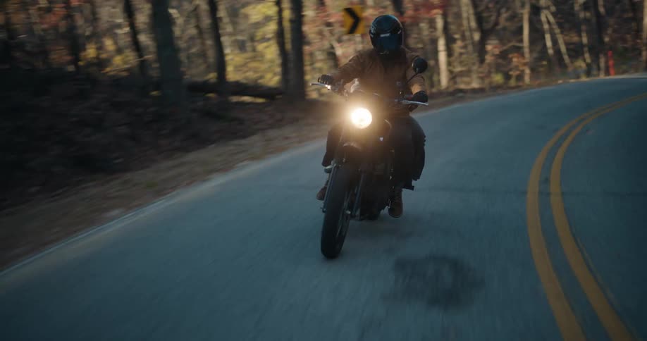 Man riding a motorcycle with a passenger on a road through the forest as a truck follows behind