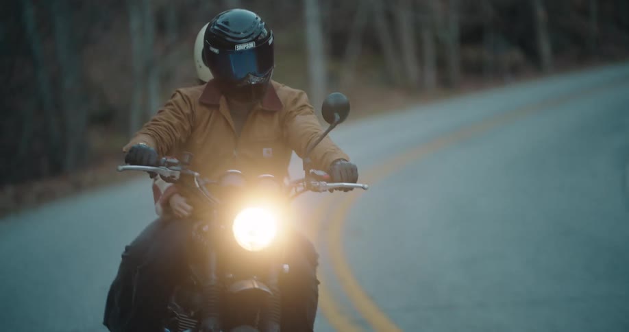A man in a brown jacket and black helmet rides a motorcycle along a winding road through a forest at dusk while a female passenger sits behind him