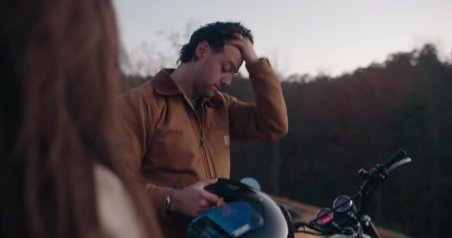 A man removes a black motorcycle helmet and runs a hand through his hair while standing near a motorcycle he then places the helmet down on the seat of the bike as a woman watches from the foreground