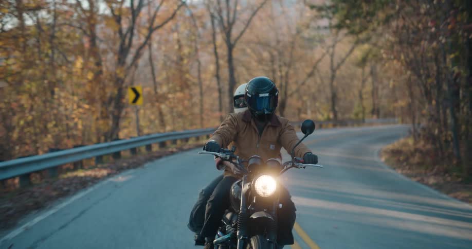 A man and woman ride a black motorcycle along a winding mountain road during autumn as sunlight flares through the trees and cars passes by