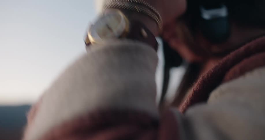 A woman wearing a striped wool sweater and several bracelets takes off a helmet of her head against a blurred backdrop of hills at dusk