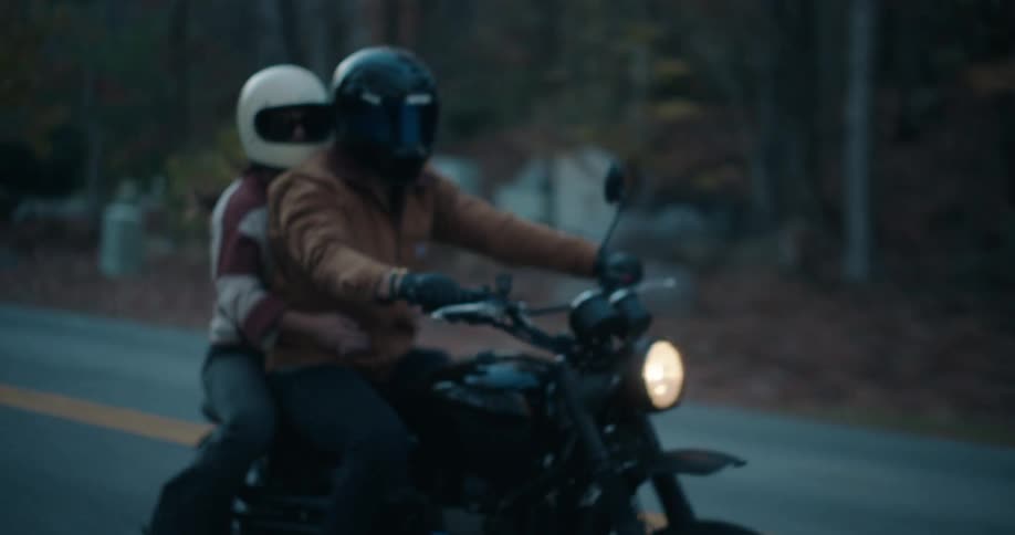 Man rides a motorcycle on a forested mountain road as his passenger holds on and they pass cars