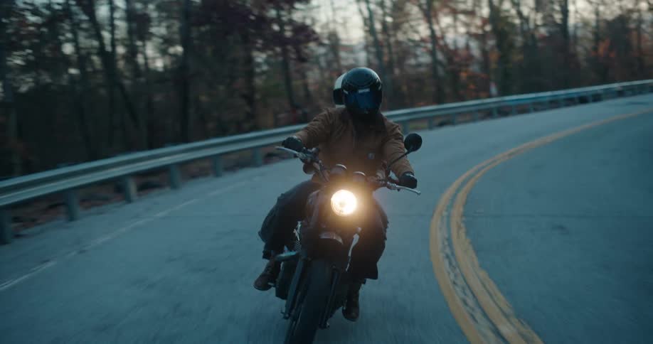 A motorcyclist and a passenger ride a black bike along a winding mountain road at dusk leaning into a sharp righthand curve while following the double yellow lines past a metal guardrail and autumn trees