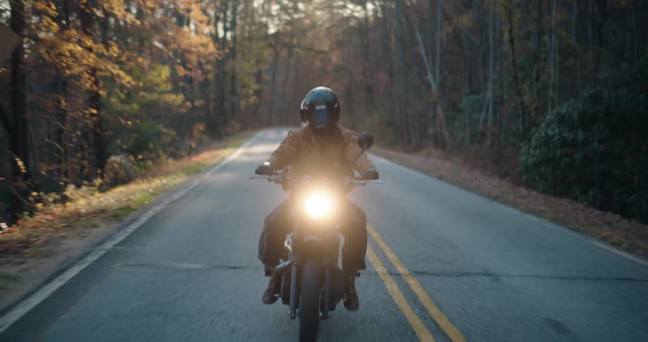 A man and woman ride a motorcycle together down a winding twolane road through a dense autumn forest as the camera pulls back from their faces to a lowangle shot of the front wheel and pavement