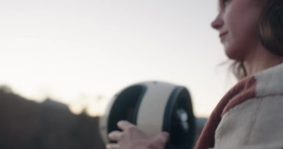 A woman wearing a patterned sweater and a leatherstrapped watch removes a white motorcycle helmet as she takes it off she reveals long dark hair against a softfocus background of hills at dusk
