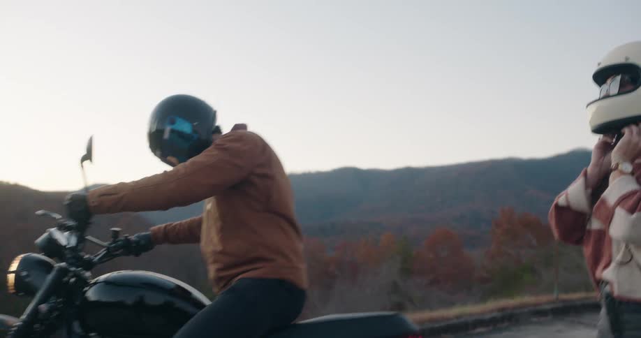 A helmeted motorcyclist in a brown jacket leans forward to steady a sleek black bike on a quiet mountain overlook at dusk while a second rider in a white helmet and striped jacket stands just behind rolling hills and autumncolored trees are in the back
