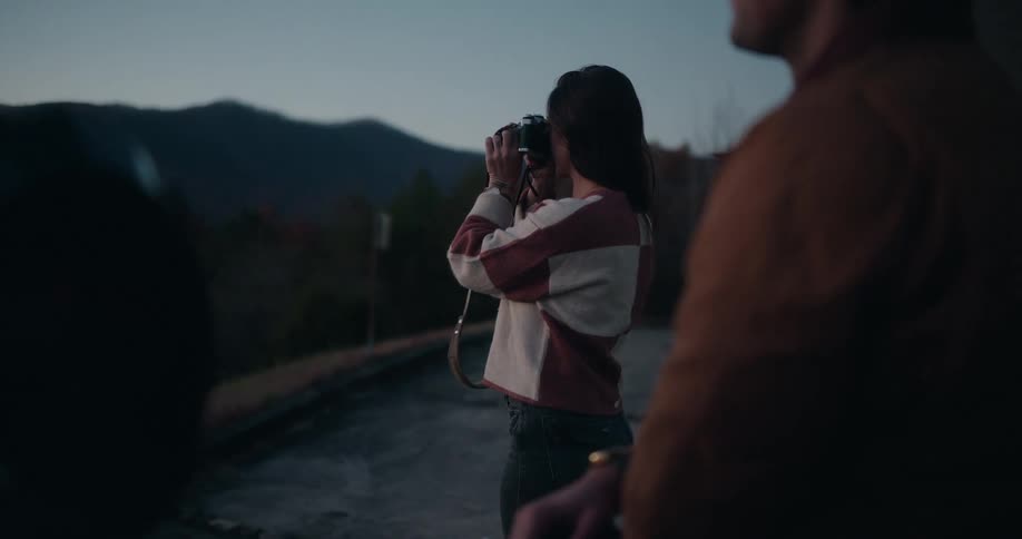 Person photographs a mountainous landscape at dusk while another stands nearby in the background