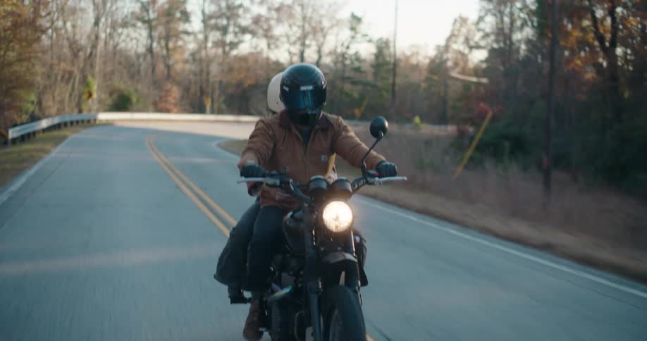 Two people ride a motorcycle on a countryside freeway at midday both wearing helmets with the driver in a brown jacket