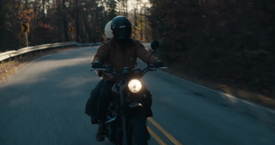 Headlight glows on a motorcycle traveling with a passenger on a road through the forest