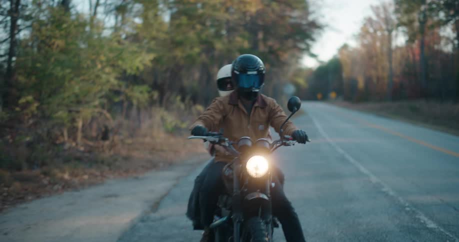 A man in a brown jacket rides a motorcycle with a passenger on a paved rural road lined with trees during an autumn day