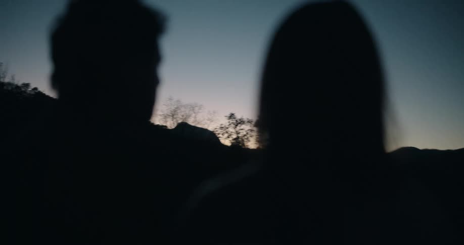The silhouettes of a couple are shown in soft focus as they look at one another against a dark twilight sky distant mountain ridges and sparse trees are visible in the background under the fading glow of dusk