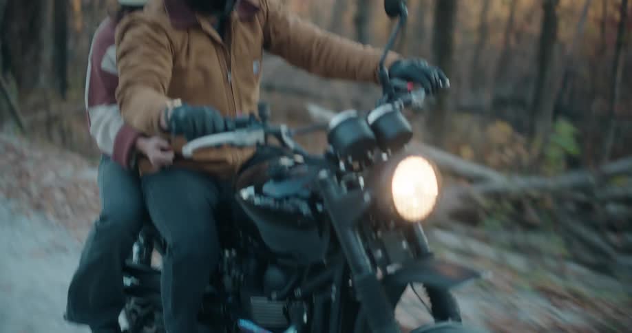 Two helmeted riders travel together on a motorcycle along a narrow forest road the driver in a brown jacket gripping the handlebars while the passenger sits close behind