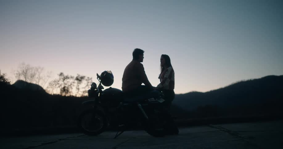 Couple sits beside a motorcycle conversing in a setting surrounded by mountain silhouettes
