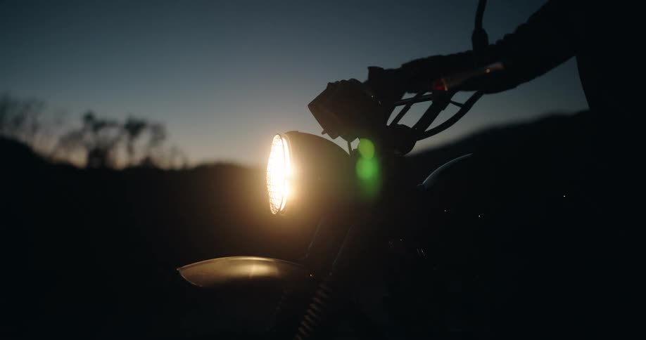 A motorcycles headlight glows warmly in the fading blue of dusk its circular beam and silhouetted handlebars cutting through the darkness
