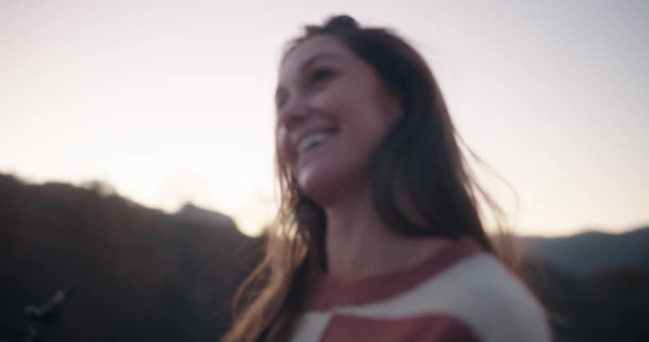 Two people smile while talking outdoors at dusk with a motorcycle and blurred natural landscape in the background