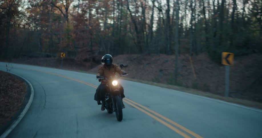 Headlight glows on a motorcycle traveling with a passenger on a road through forested mountains