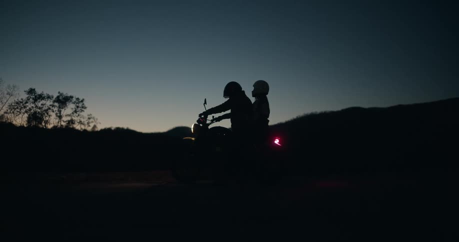 Two people on a motorcycle appear in silhouette at dusk on a road surrounded by hills