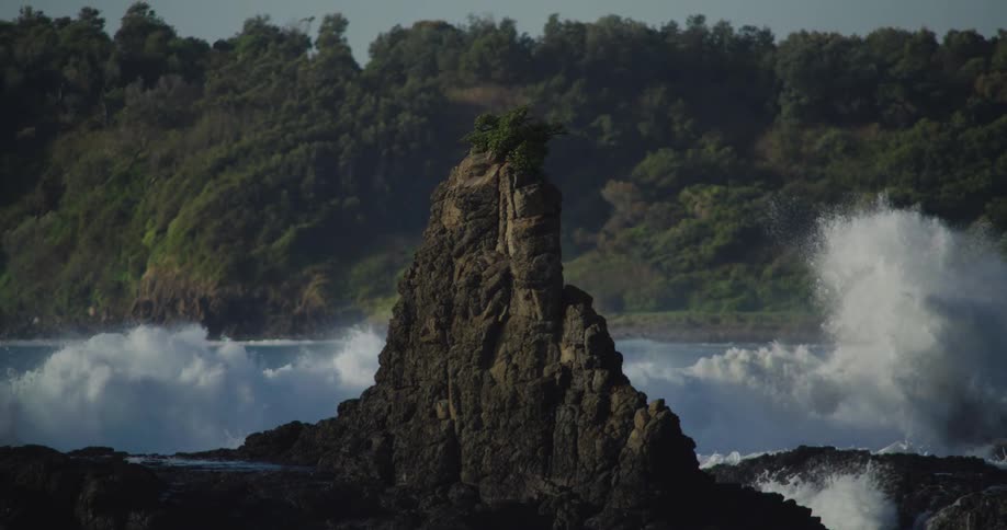 Forested mountains along the coast as waves splash into a rock formation in the water