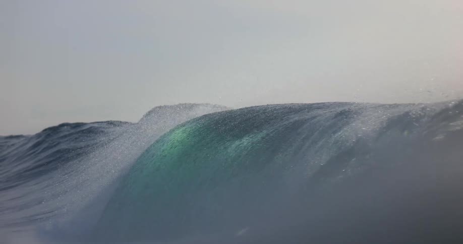 A closeup shot captures a turquoise ocean wave as it crests and begins to break sending sea spray into the air before the camera is submerged in white mist and water