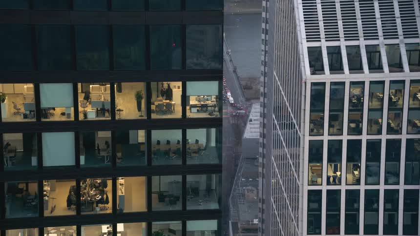 Corporate buildings display illuminated offices seen through glass facades in a dense urban area