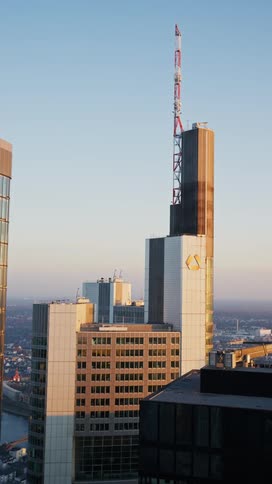 A tall skyscraper with a yellow logo and a redandwhite antenna stands prominently against a clear sky during sunset surrounded by other office buildings