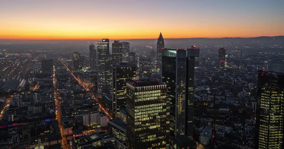 An aerial twilight view shows a city skyline as city lights flicker on and the sun sets behind distant hills casting a golden glow over the dense cluster of skyscrapers and the illuminated streets stretching into the urban horizon