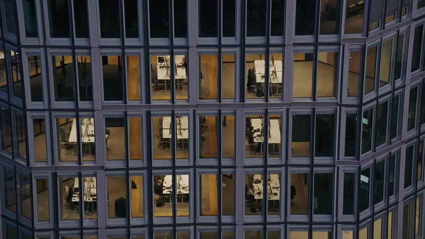 Through the grid of a modern glass office building at dusk rows of illuminated workspaces reveal people seated at desks creating a quiet repetitive pattern