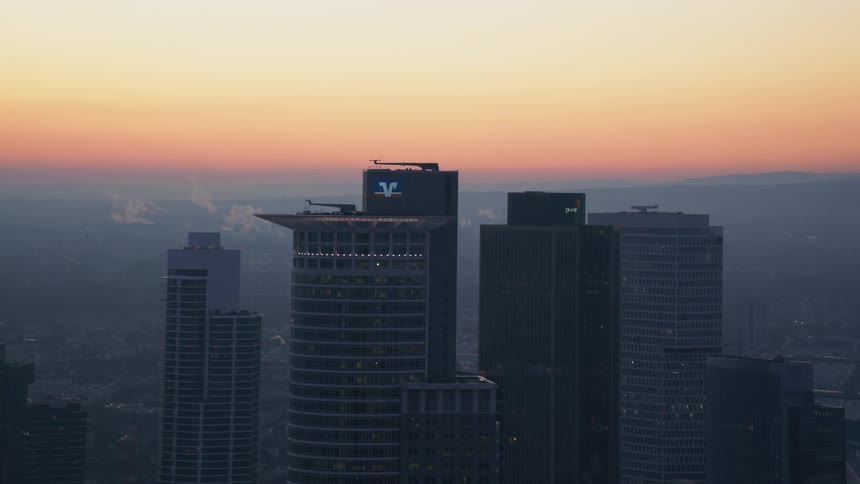 A wide elevated view captures a city skyline at dawn showcasing several prominent skyscrapers against an orange and hazy purple horizon while thin trails of steam rise from the rooftops