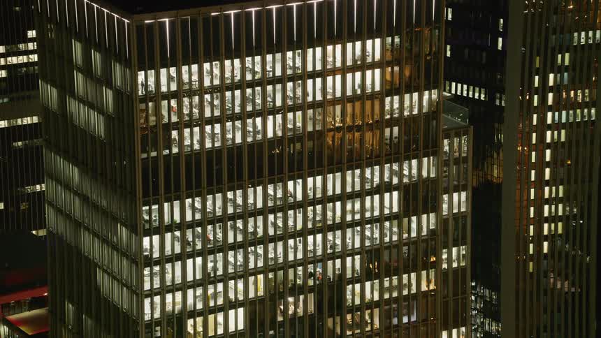 A nighttime highangle shot shows a glowing glass skyscraper where several floors of a modern office are visible featuring rows of workstations and lit ceilings against the dark urban background of a city