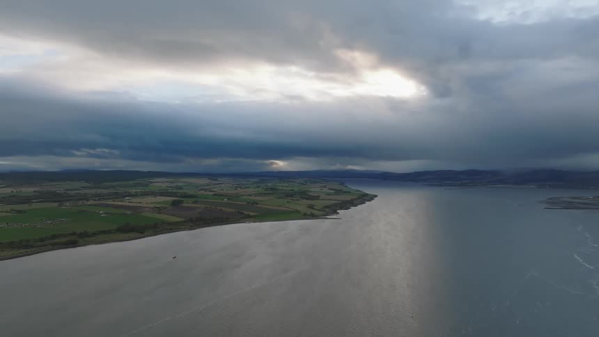 A broad silvery body of water bordered by patchwork green farmland and distant hills as heavy layered storm clouds part slightly to let pale light spill across the landscape