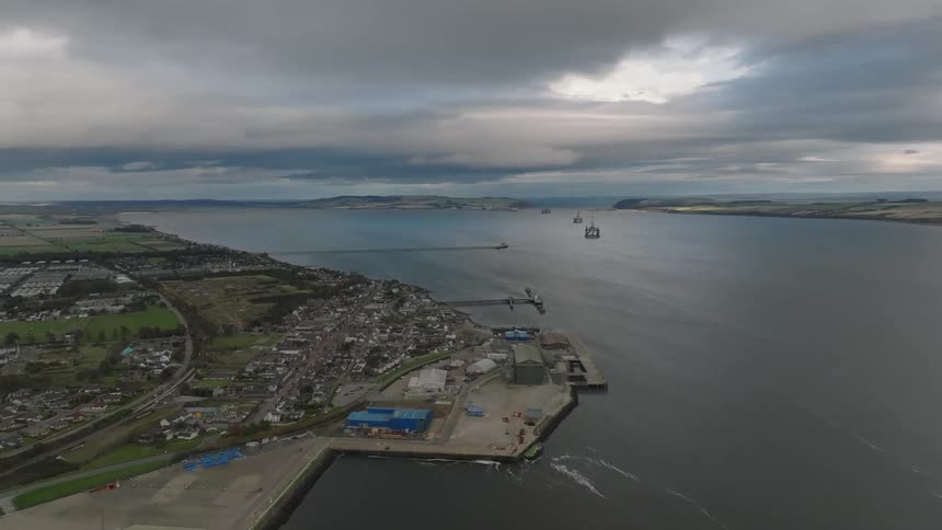 A wide view of a coastal town and its industrial port along a deep bay under a heavy overcast sky while several large oil platforms are stationed in the distance near the mouth of the waterway