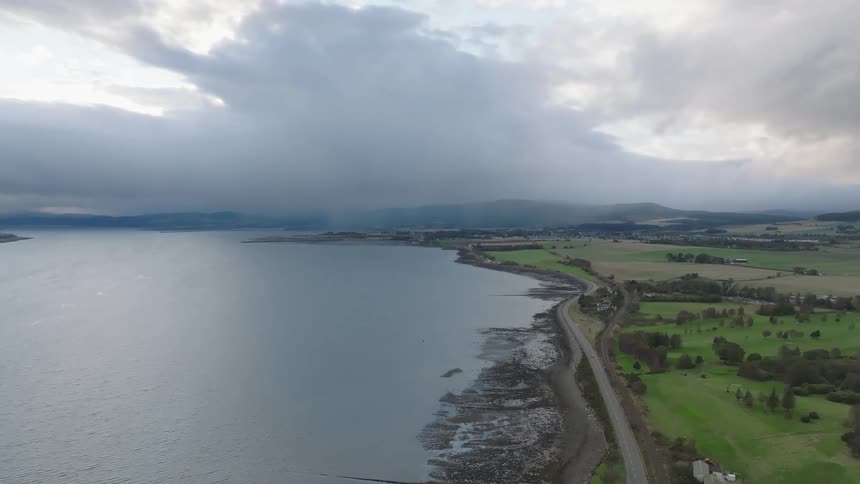 Extended coastline with a coastal road running alongside rural areas and green fields under a cloudy sky