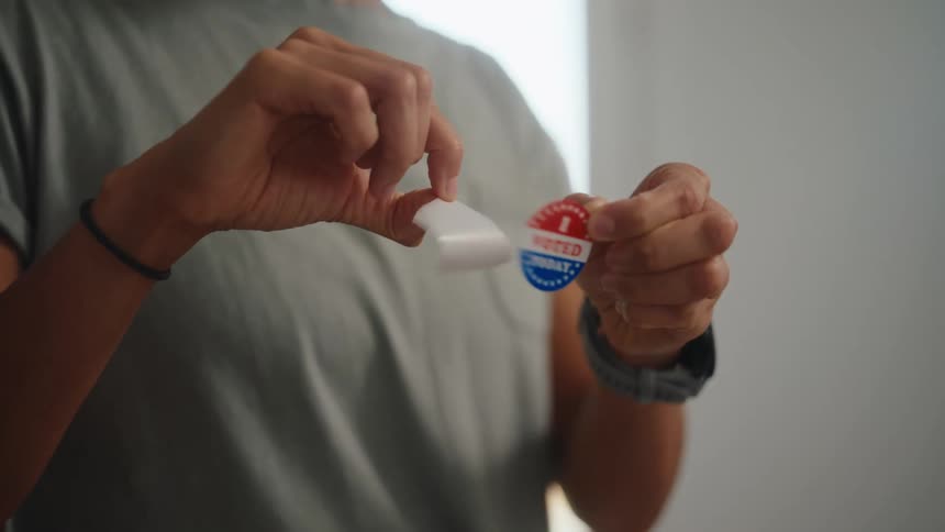 A persons hands are shown carefully peeling a circular i voted today sticker off of its white paper backingonce the red white and blue sticker is fully removed the person holds it up while still grasping the empty adhesive strip in their other ha