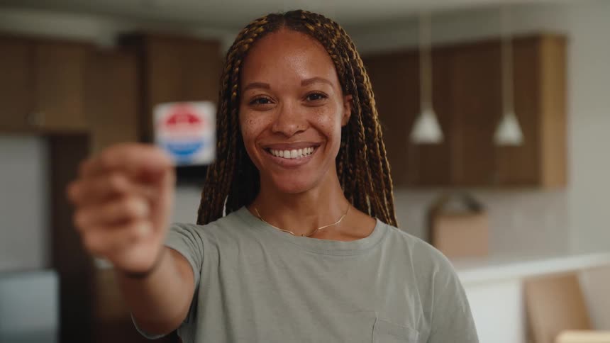 A smiling woman with long blonde and brown braids stands in a kitchen and holds up an i voted sticker toward the camerathe scene captures her looking directly at the viewer with a joyful expression celebrating the completion of her civic duty in a