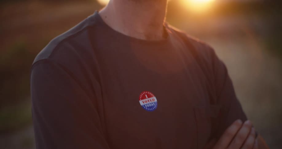 Man with crossed arms wears a dark shirt with a voting sticker on the chest under natural outdoor lighting