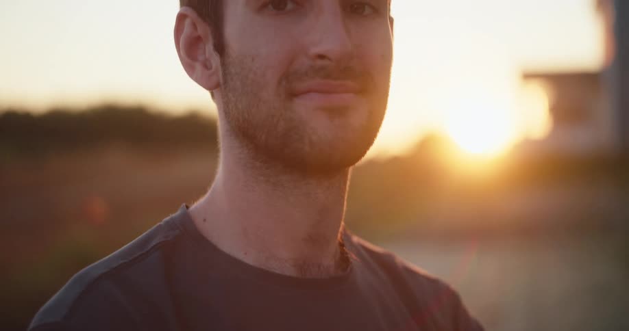 A young man wearing a black tshirt and an i voted today badge smiles with the setting sun glowing behind him