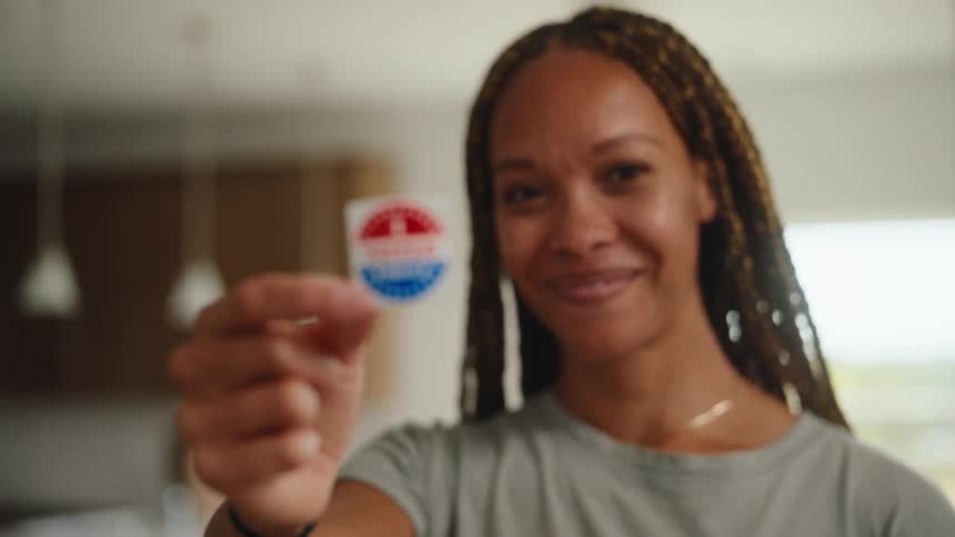 A smiling woman with braided hair stands in a kitchen and holds up an i voted today sticker toward the camera shifting the focus from her face to the circular red white and blue sticker held between her fingers