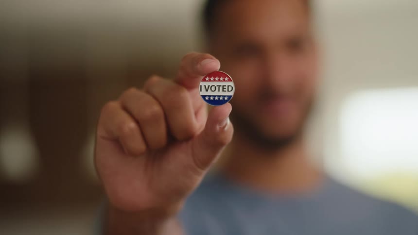 A person holds a small circular i voted sticker toward the camera while smiling the sticker features red and white stripes with blue stars set against a blurred indoor background