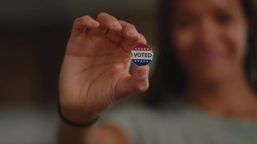 Hand in the foreground holds an i voted button while a person appears blurred in the background
