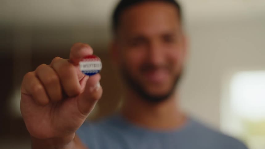 A man looks into the camera with a warm smile as he slowly brings an i voted sticker into clear focusthe circular sticker is red white and blue and he holds it steadily toward the viewer to display it proudly