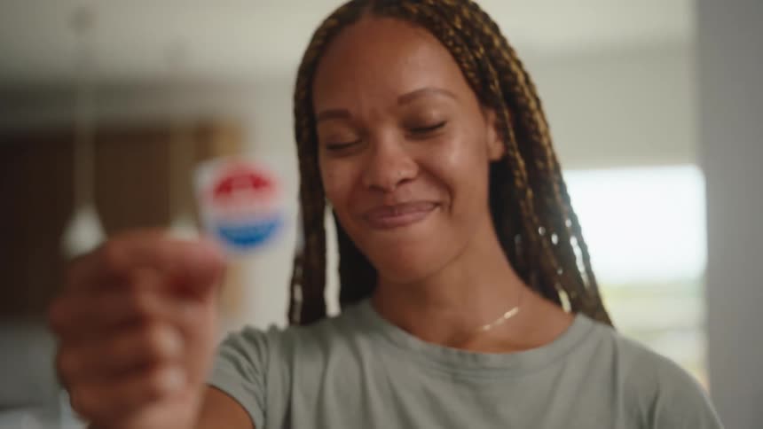 A smiling woman with long braids holds up an i voted today sticker toward the camera in a brightly lit kitchen