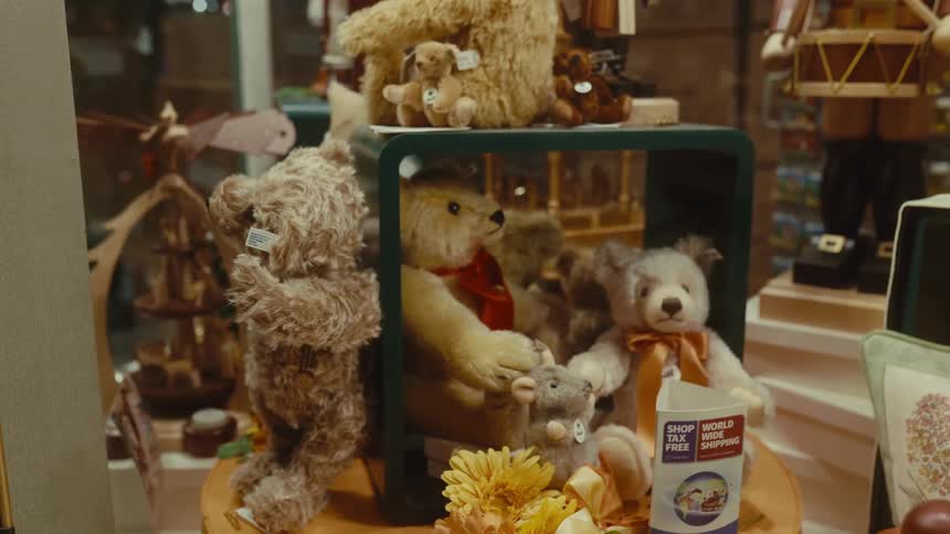 A shop window display features an array of classic teddy bears and holidaythemed figurines arranged on wooden stands the camera slowly pans across the festive scene highlighting a large nutcracker and a sign for taxfree world shipping