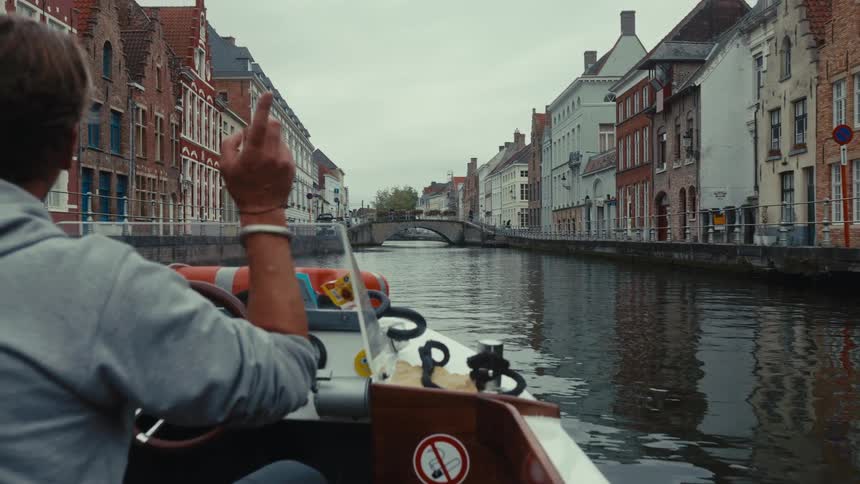 Person steers a boat along an urban canal lined with historic buildings and a bridge in the distance