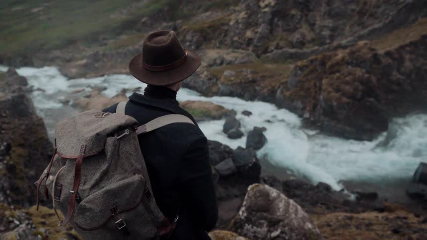 A lone traveler standing on a cliff overlooking a rushing river in a rocky mountain landscape