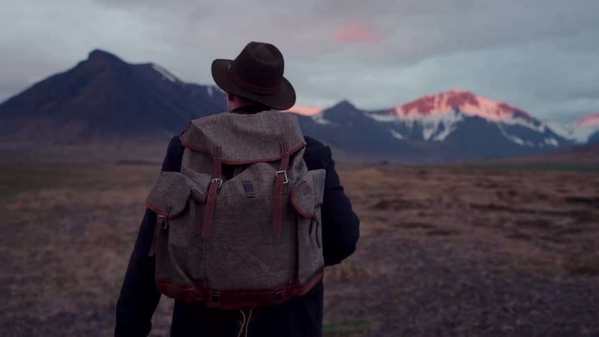 Person wearing a backpack and hat looks toward an open landscape with mountains in the background under a partly cloudy sky