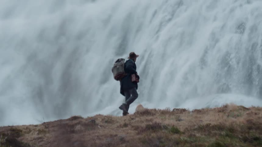 A man wearing a hat and carrying a large backpack walks along a rocky ridge a massive powerful waterfall cascades down the cliffside directly behind him as he moves