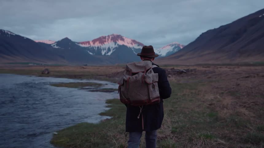 Person wearing a backpack walks along a riverbank in a mountainous landscape with snowcapped peaks in the background