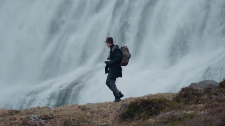 A man wearing a hat and backpack walks across a grassy ridge in front of a massive misty waterfall he carries a camera around his neck as he moves from right to left against the backdrop of the cascading water